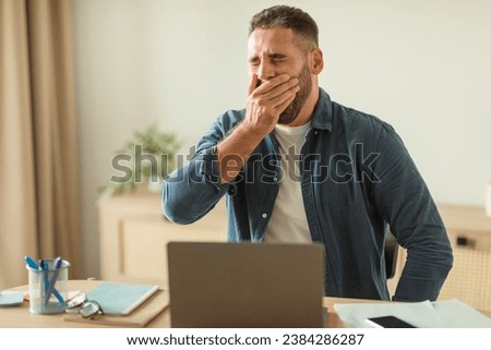 Tired middle aged man yawning while sitting at laptop in modern office. Bored and sleepy employee man suffering after overwork at workplace in early morning. Professional burnout, exhaustion