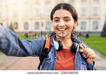 Portrait of cheerful pretty young indian woman student taking selfie outdoors at university campus, wearing casual outfit, wireless headphones, carrying backpack, blogging after classes
