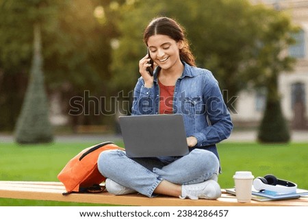 Part-time job, freelance for students. Positive beautiful young indian woman using computer laptop and talking on phone with employer, chilling on lawn at public park, doing homework, free space Royalty-Free Stock Photo #2384286457
