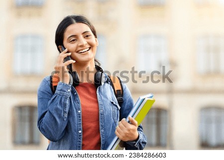 Joyful attractive young indian student strolling through the college grounds, chatting on her mobile with a mate, organizing meet-ups, with textbooks in grasp and a rucksack on her back, blank space. Royalty-Free Stock Photo #2384286603