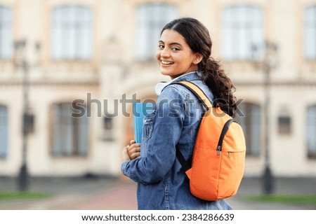 A radiant young Indian scholar with her academic books and bag, posing outside her educational institution, offering a warm smile to the lens. Space for caption, emphasizing education. Royalty-Free Stock Photo #2384286639