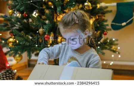 Excited curious little girl smiling, opening christmas gifts. Beautifully decorated christmas tree and house with lights and lantern.