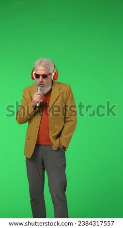 Portrait of senior stylish hipster on Chroma key green screen background, man in headphones sings in microphone. Advertising area, workspace mockup. Vertical photo.