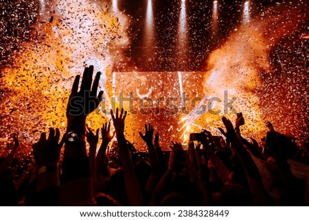 Bright lights of a concert show, fans' hands, vibrant lights, and confetti. Red and orange hues illuminate the cheering crowd with hands raised high. Royalty-Free Stock Photo #2384328449