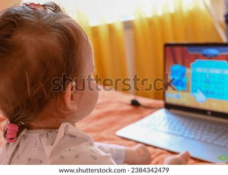 The little girl is sitting on the bed and watching a cartoon on the laptop. The baby is watching cartoons on the computer.