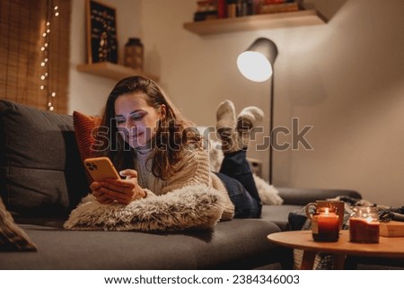 A young woman spends a cozy winter evening at home lying on the sofa looking at her smartphone. Winter holidays, Christmas and online surfing 
