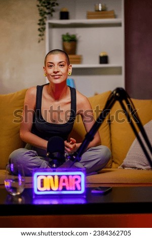 Portrait of a bald woman talking about important topics live on air while recording a podcast episode in a cozy studio on a yellow sofa. Content creator or radio show concept. Looking into the camera.
