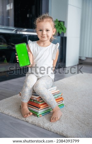 Little girl is sitting on stack of children's books and holding a smartphone with a chromakey. High quality photo
