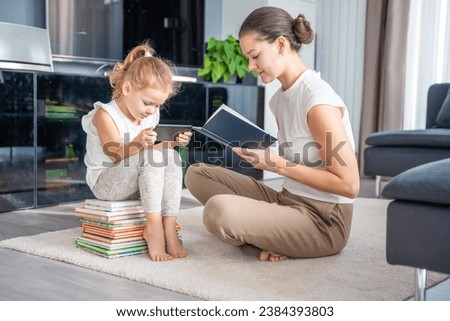 Little girl sits on a stack of children's books and uses her smartphone while mother reads a book. High quality photo
