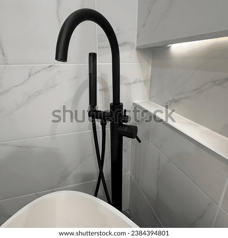 A modern black shower faucet and handheld showerhead, set against a background of white marble tiles with subtle grey veining and a lit recessed shelf.