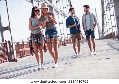Cheerful young people are talking and smiling while walking in the city, girls are using a smart phone