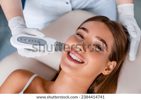 Smiling caucasian female on ultrasound facial beauty treatment. Cosmetologist doing facial cleaning peeling in beauty salon for anti-age rejuvenation pampering effect