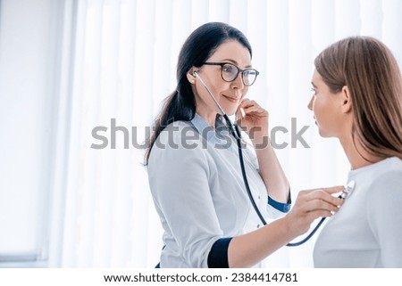 Female doctor listening to patients chest with stethoscope at the hospital, checking on lungs illnesses diseases. General practitioner listening to patient`s heart beat, cardiology concept Royalty-Free Stock Photo #2384414781