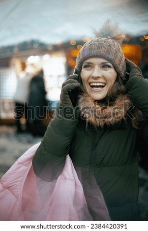 smiling modern female in green coat and brown hat at the winter fair in the city talking on a smartphone.