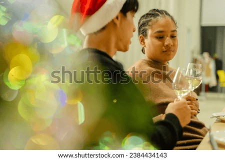couple man and woman holding champagne glass and sitting at dinner party christmas and new year. Cheers with wine glasses in beautiful meeting of family gathering. Home decor with Christmas tree. 