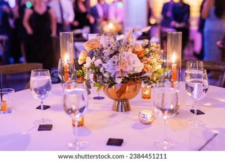 Wedding reception with purple uplighting, a close-up of a table with a floral arrangement and water glasses on a white table. Royalty-Free Stock Photo #2384451211