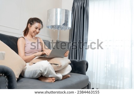 Video call, a young Asian woman communicates via online video long distance, looking at the digital laptop screen, happily greeted with a friend in her house.