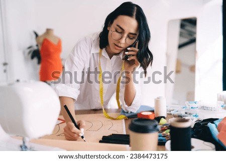 Craftswoman drawing draft and chatting on smartphone in workroom