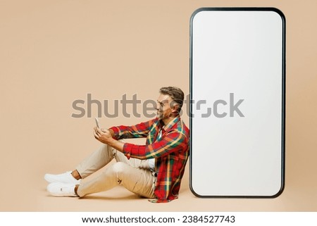 Full body side view adult man wearing red shirt white t-shirt casual clothes sit near big huge blank screen mobile cell phone smartphone with area use device isolated on plain beige color background