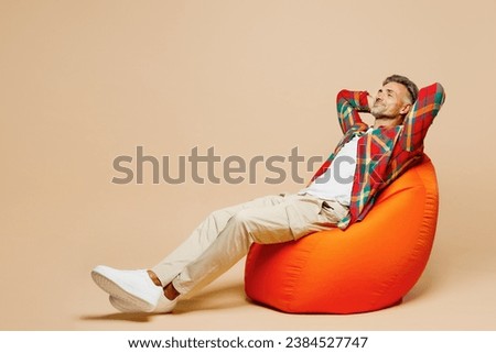 Full body side view adult man he wear red shirt white t-shirt casual clothes sit in bag chair hold hands behind neck isolated on plain pastel light beige background studio portrait. Lifestyle concept