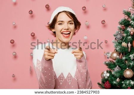 Merry young woman wears white sweater hat posing point index finger camera on you motivating encourage isolated on plain pastel pink background. Happy New Year celebration Christmas holiday concept