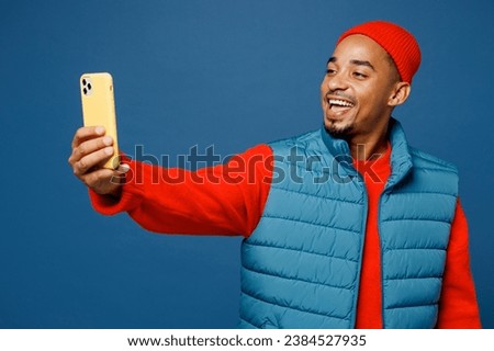 Young man of African American ethnicity wear padded vest red hat doing selfie shot on mobile cell phone post photo on social network isolated on plain dark royal navy blue background studio portrait