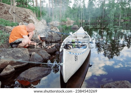 A man prepares breakfast on a small camping stove next to a metal canoe by a lake in Sweden Royalty-Free Stock Photo #2384543269