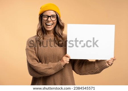Young  happy attractive woman winks wearing yellow hat and stylish eyeglasses holding empty placard  isolated on background, mockup. Portrait of smart smiling student with poster looking at camera Royalty-Free Stock Photo #2384545571