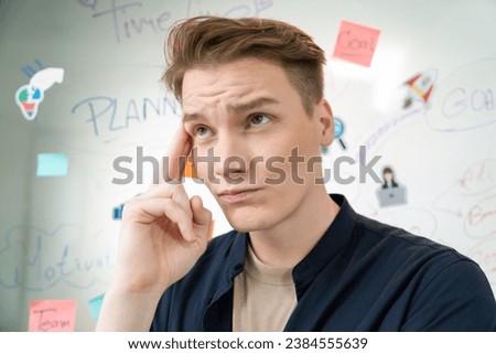 Portrait of young caucasian businessman thinking with confused face expression while standing in front of glass board with sticky notes and mind map at creative business meeting. Immaculate.