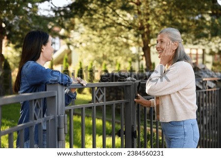 Friendly relationship with neighbours. Happy women talking near fence outdoors Royalty-Free Stock Photo #2384560231