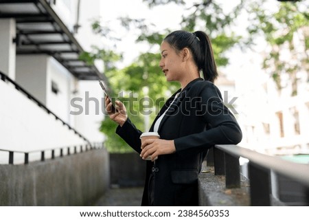 Side view image of a beautiful millennial Asian businesswoman in a formal business suit is reading messages or online blogs on her smartphone while taking a coffee break outdoors.