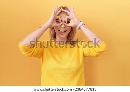 Young caucasian woman wearing yellow sweater doing ok gesture like binoculars sticking tongue out, eyes looking through fingers. crazy expression. 