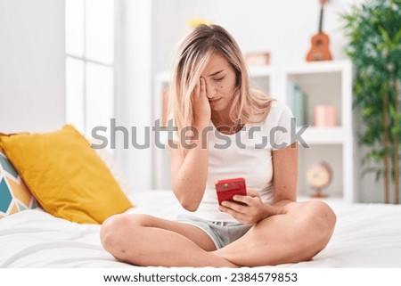 Young blonde woman using smartphone with worried expression at bedroom