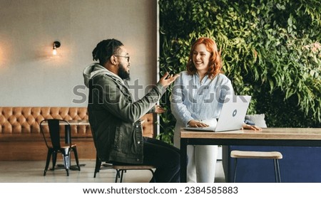 Happy colleagues collaborating in a vibrant cafe, discussing and using a laptop for a project. Professionals in the tech industry exemplify successful teamwork and remote work in a coworking space. Royalty-Free Stock Photo #2384585883