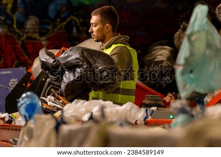 Focused male sorter with a protective vest and gloves unloads waste from a garbage bag onto a garbage sorting line