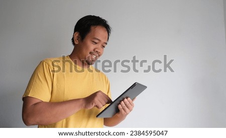 Casually dressed young Southeast Asian man smiling and using a digital tablet with gray background Royalty-Free Stock Photo #2384595047