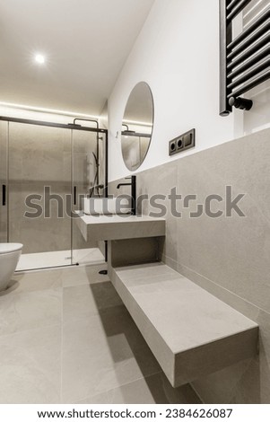 Beautiful modern bathroom with gray tiles, countertop of the same material, circular mirror, shower cabin with glass screens, wall-mounted towel radiator and black taps Royalty-Free Stock Photo #2384626087