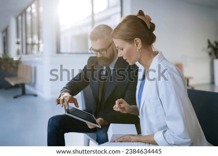 Pharmaceutical sales representative talking with female doctor in medical building, presenting new medication. Hospital director consulting with healthcare staff. Royalty-Free Stock Photo #2384634445