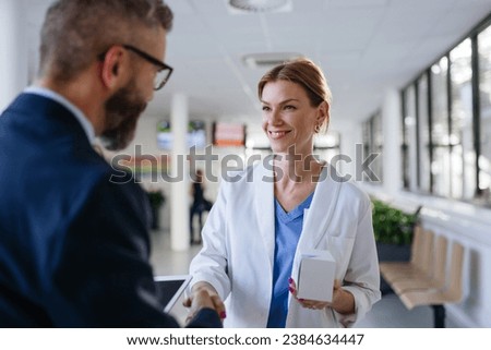 Pharmaceutical sales representative shaking hand with female doctor in medical building. Hospital director consulting with healthcare staff. Royalty-Free Stock Photo #2384634447
