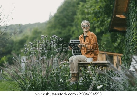 Woman in garden holding solar panel and tablet. Looking for solar panels grants, funds for homeowners. Solar energy and sustainable lifestyle for family in house. Royalty-Free Stock Photo #2384634553