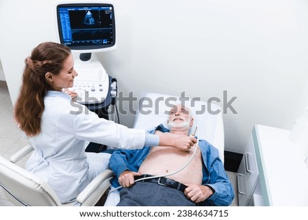 Heart ultrasound examination performed by woman doctor with smiling aged patient. Sonographer specialist. Cardiovascular diseases prevention. Heartbeat measurement Royalty-Free Stock Photo #2384634715
