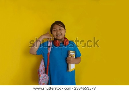 Adorable little girl holding books, wearing backpack and headphones on neck make phone gesture over yellow background