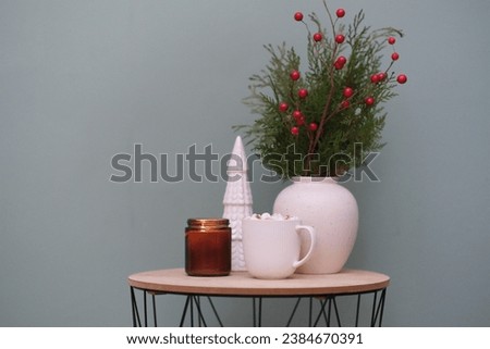 Christmas home decorations - burning candle, cup of cocao with marshmellow, ceramic christmas tree, fir branches in vase on the coffee table on green background.