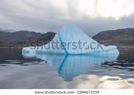 Large iceberg in Nanortalik Greenland with large blue clear water stripe and many creases and crevasses iseen with blue tinge to ice and good reflection in still water and against overcast sky Royalty-Free Stock Photo #2384673159