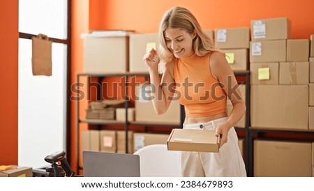 Young blonde woman ecommerce business worker using laptop holding package celebrating at office