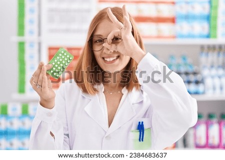 Young redhead woman working at pharmacy drugstore holding birth control pills smiling happy doing ok sign with hand on eye looking through fingers 