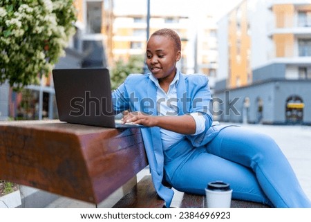 Beautiful young girl in business attire sitting on a bench in front of the office, drinking coffee and using a laptop to catch up on unanswered emails