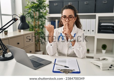 Young hispanic woman wearing doctor uniform and stethoscope asking to be quiet with finger on lips pointing with hand to the side. silence and secret concept. 