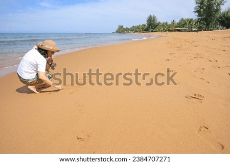 A woman sits holding a camera and wearing a hat writing the letters L O V E in the sand on a holiday morning. A young female tourist happily draws "love" on the sand at the beach while relaxing.  Royalty-Free Stock Photo #2384707271
