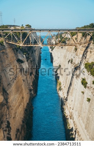 Corinth Canal. Pictured is an old bridge over the Corinth canal and a ship sails in the canal in the distance. The channel is deep and built in the rock. The water in the canal is dark blue.